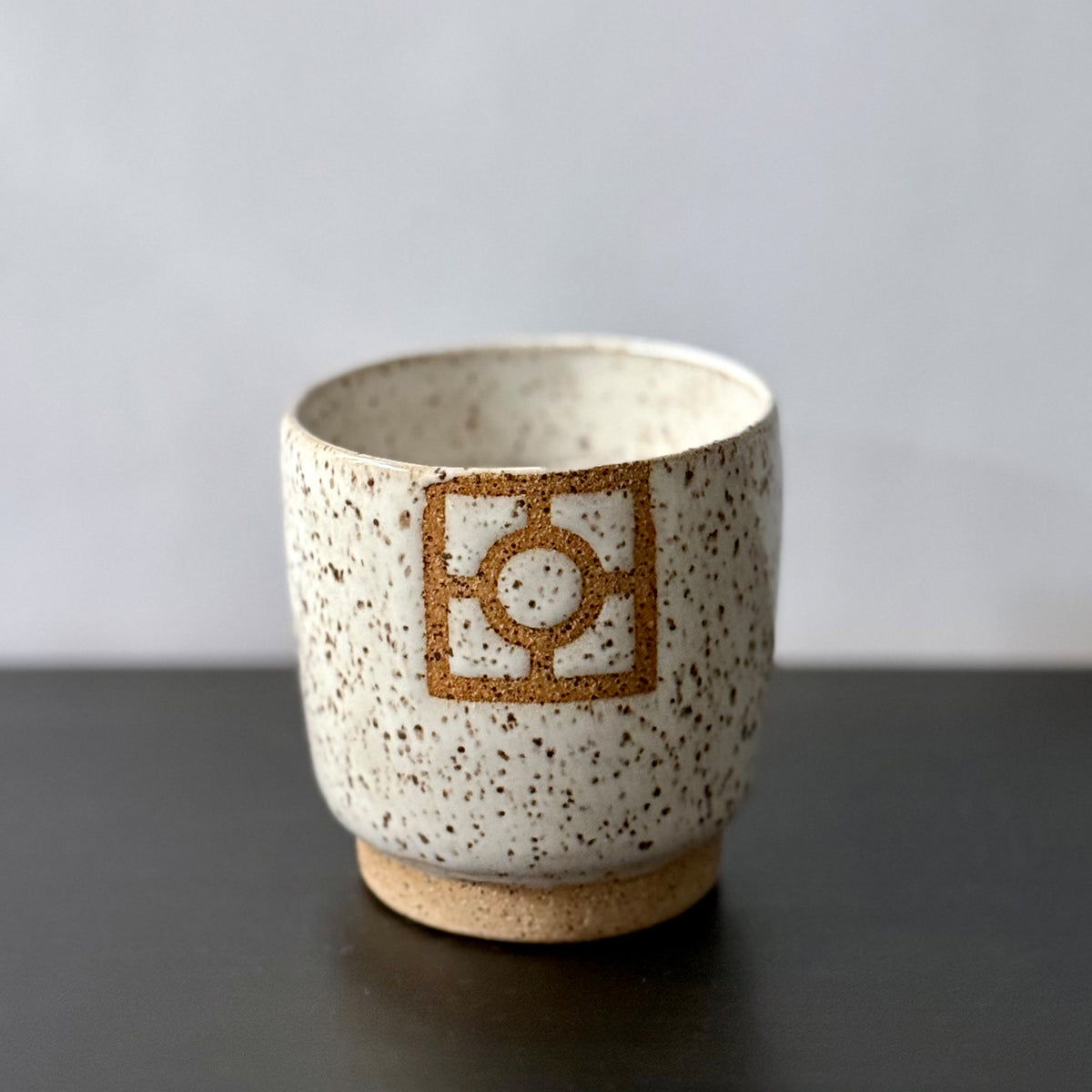 Handmade ceramic tumbler with a rounded shape, speckled white glaze, exposed stoneware foot, and a square geometric breeze-block motif on the front with a circular center detail.