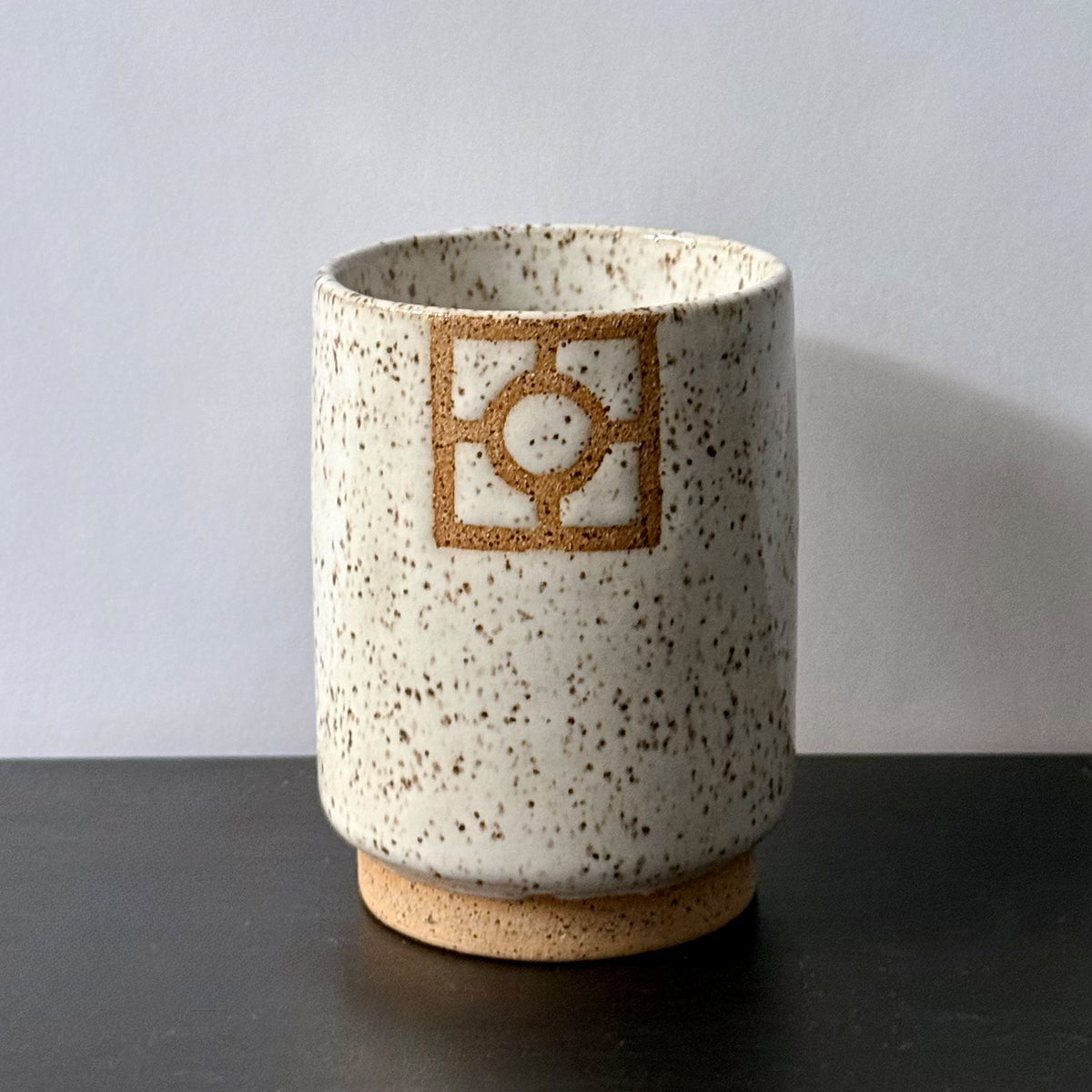 Handmade ceramic tumbler with a tall cylindrical shape, speckled white glaze, exposed stoneware foot, and a square geometric breeze-block motif on the front with a circular center detail.
