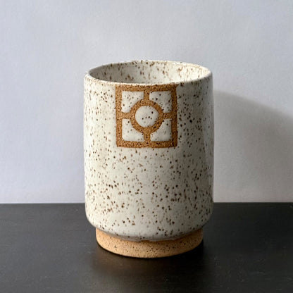 Handmade ceramic tumbler with a tall cylindrical shape, speckled white glaze, exposed stoneware foot, and a square geometric breeze-block motif on the front with a circular center detail.