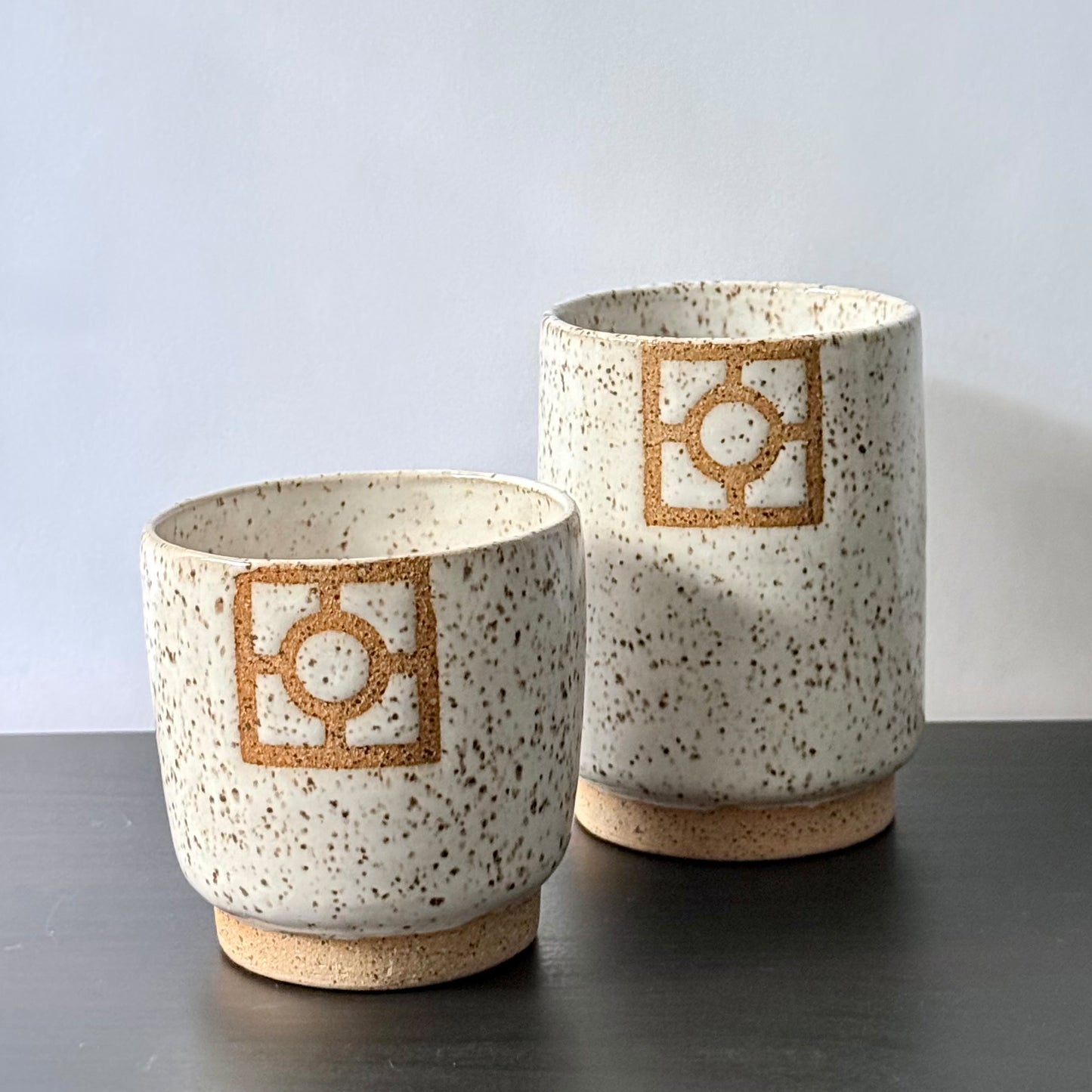 Two handmade ceramic tumblers with a speckled white glaze, exposed stoneware foot rings, and square geometric breeze-block motifs on the front. One tumbler is slightly taller than the other, with both featuring warm natural clay details against the light glaze.