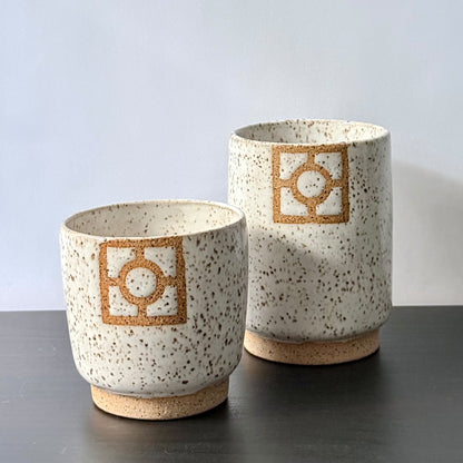 Two handmade ceramic tumblers with a speckled white glaze, exposed stoneware foot rings, and square geometric breeze-block motifs on the front. One tumbler is slightly taller than the other, with both featuring warm natural clay details against the light glaze.