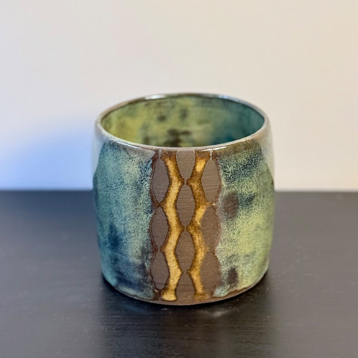 Handmade ceramic tumbler with a rounded shape and mottled glaze in mossy green, teal, blue, and warm ochre. A vertical geometric breeze-block motif on the front reveals areas of raw stoneware, creating a bold mid-century modern contrast.