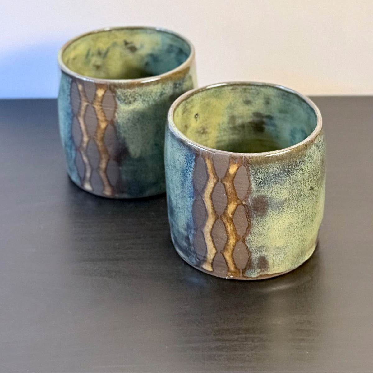 Handmade ceramic tumblers with a rounded shape and mottled glaze in mossy green, teal, blue, and warm ochre. A vertical geometric breeze-block motif on the front reveals areas of raw stoneware, creating a bold mid-century modern contrast.