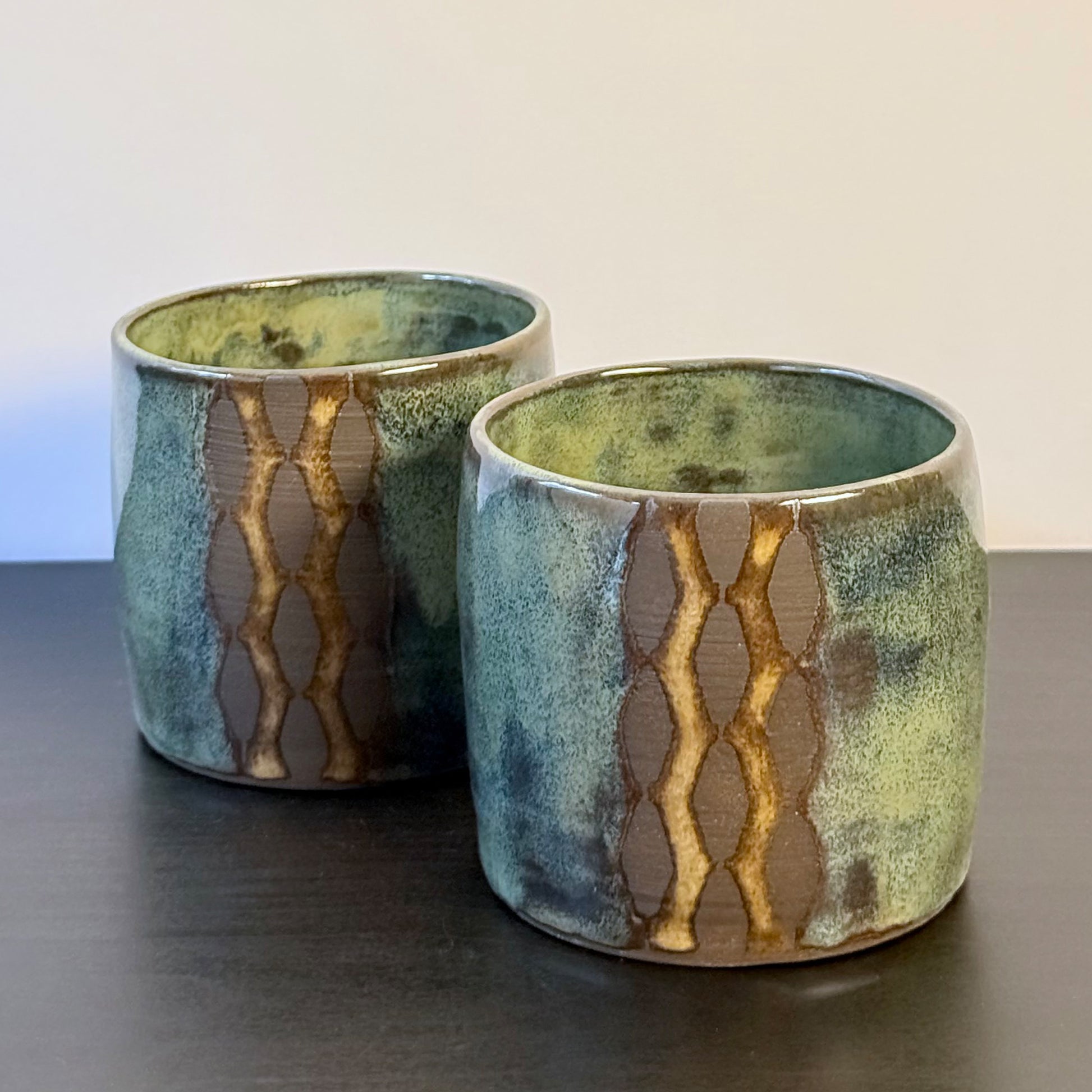 Handmade ceramic tumblers with a rounded shape and mottled glaze in mossy green, teal, blue, and warm ochre. A vertical geometric breeze-block motif on the front reveals areas of raw stoneware, creating a bold mid-century modern contrast.