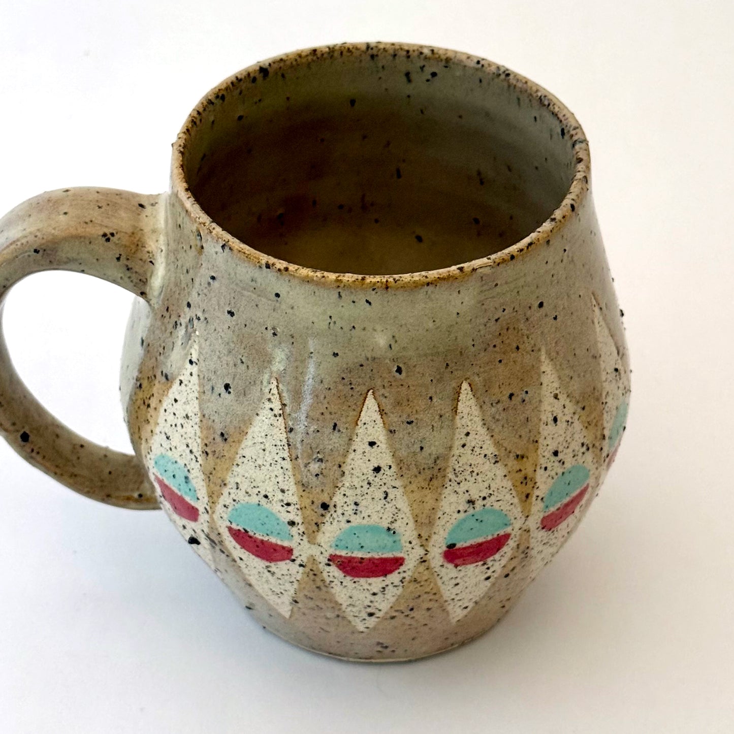 A bell-shaped, speckled tan stoneware mug featuring a pattern of cream diamonds. Each diamond contains a circular motif split into teal and poppy red. The mug has a large, rounded handle and visible dark mineral flecks.