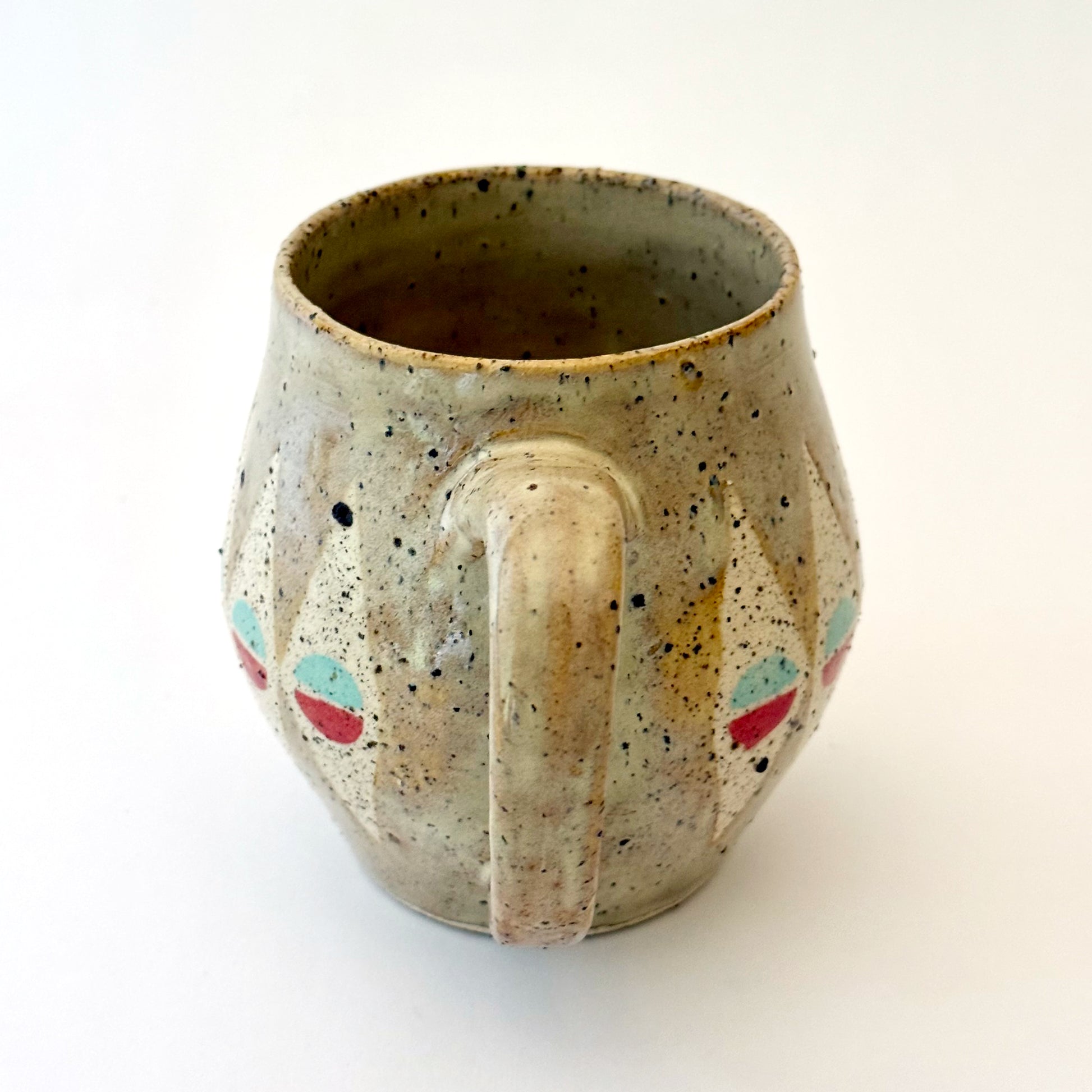 A bell-shaped, speckled tan stoneware mug featuring a pattern of cream diamonds. Each diamond contains a circular motif split into teal and poppy red. The mug has a large, rounded handle and visible dark mineral flecks.