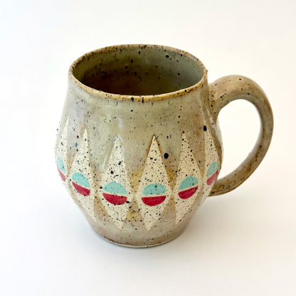 A bell-shaped, speckled tan stoneware mug featuring a pattern of cream diamonds. Each diamond contains a circular motif split into teal and poppy red. The mug has a large, rounded handle and visible dark mineral flecks.