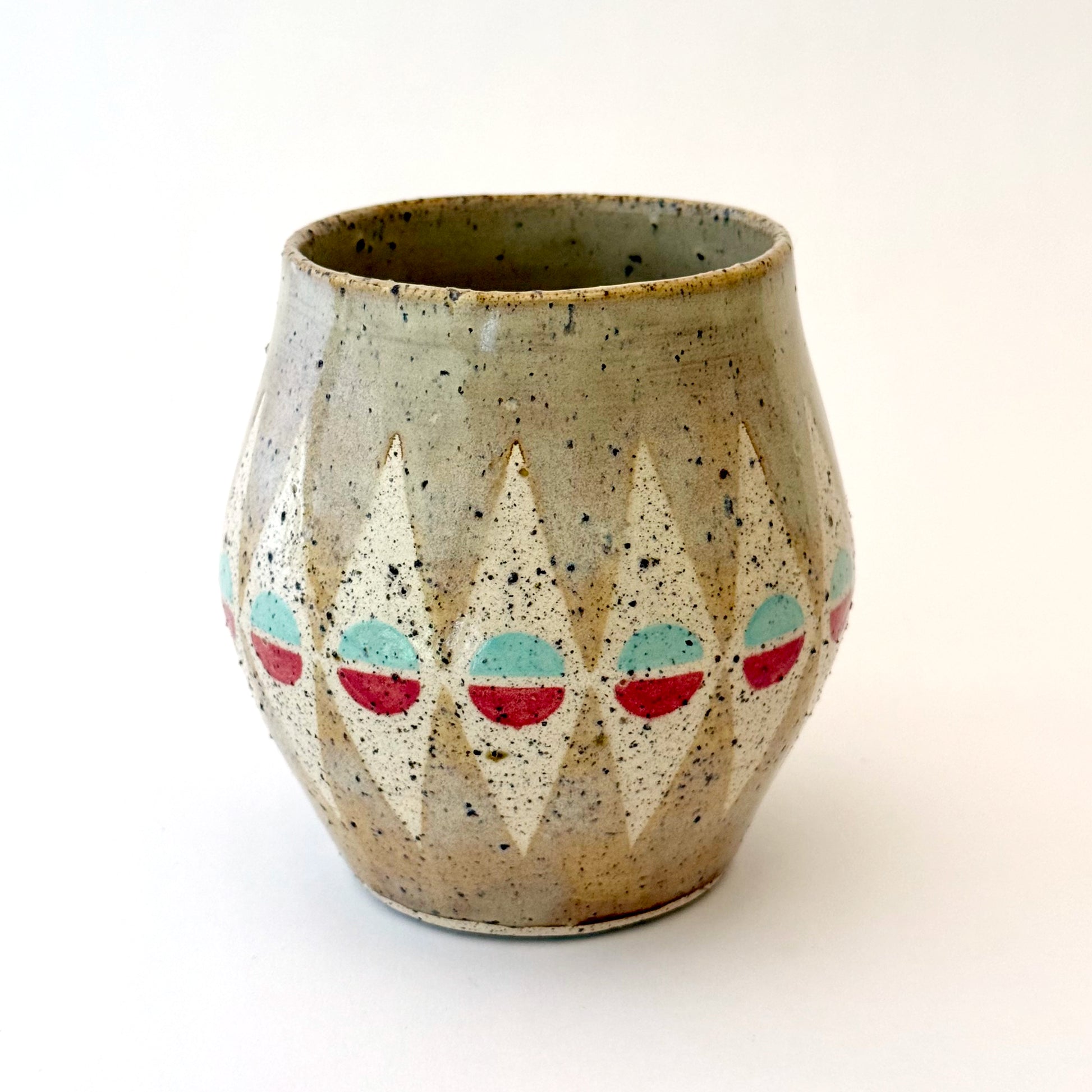 A bell-shaped, speckled tan stoneware mug featuring a pattern of cream diamonds. Each diamond contains a circular motif split into teal and poppy red. The mug has a large, rounded handle and visible dark mineral flecks.