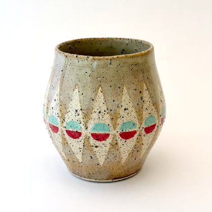 A bell-shaped, speckled tan stoneware mug featuring a pattern of cream diamonds. Each diamond contains a circular motif split into teal and poppy red. The mug has a large, rounded handle and visible dark mineral flecks.