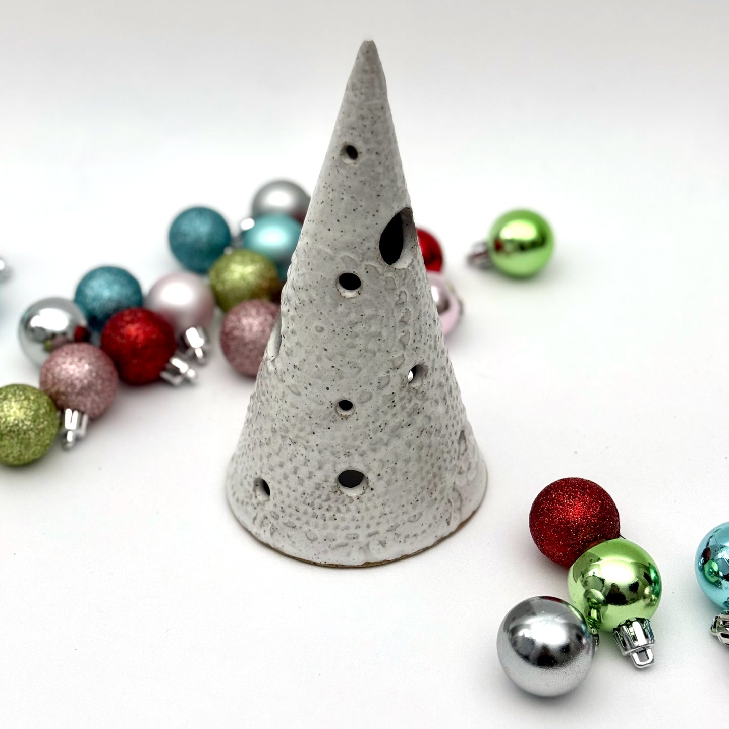 A white speckled ceramic tree-shaped luminary with light openings, styled among colorful holiday ornaments.
