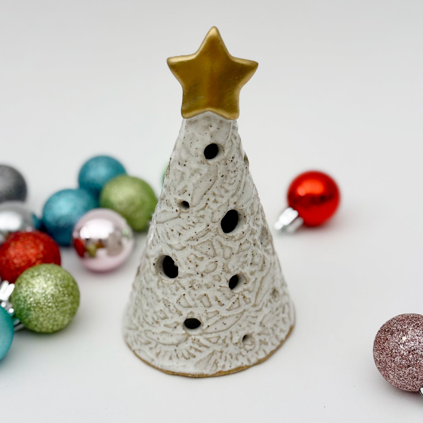 Decorative ceramic Christmas tree luminary with a gold star on top, surrounded by colorful ornaments on a light background.