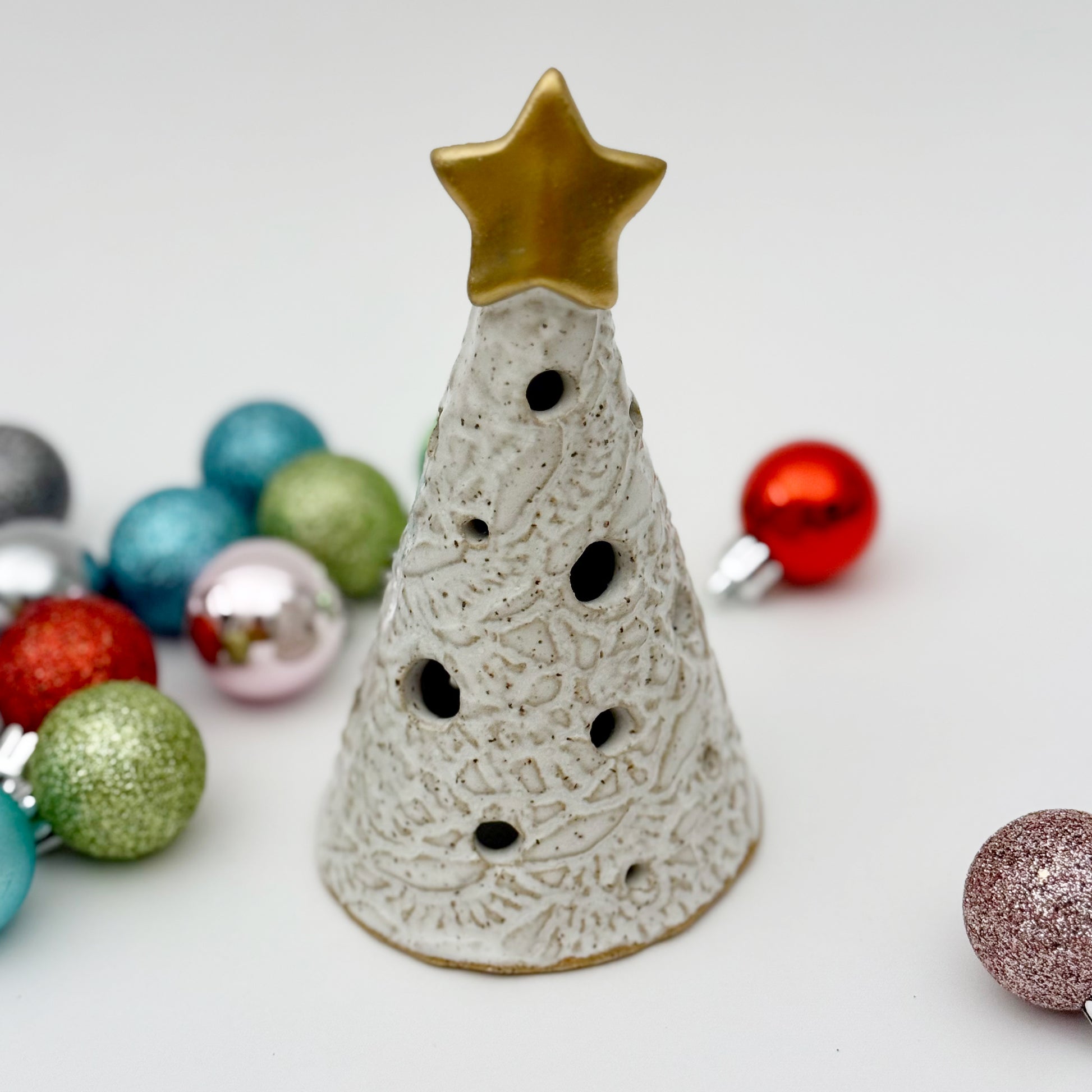 Decorative ceramic Christmas tree luminary with a gold star on top, surrounded by colorful ornaments on a light background.
