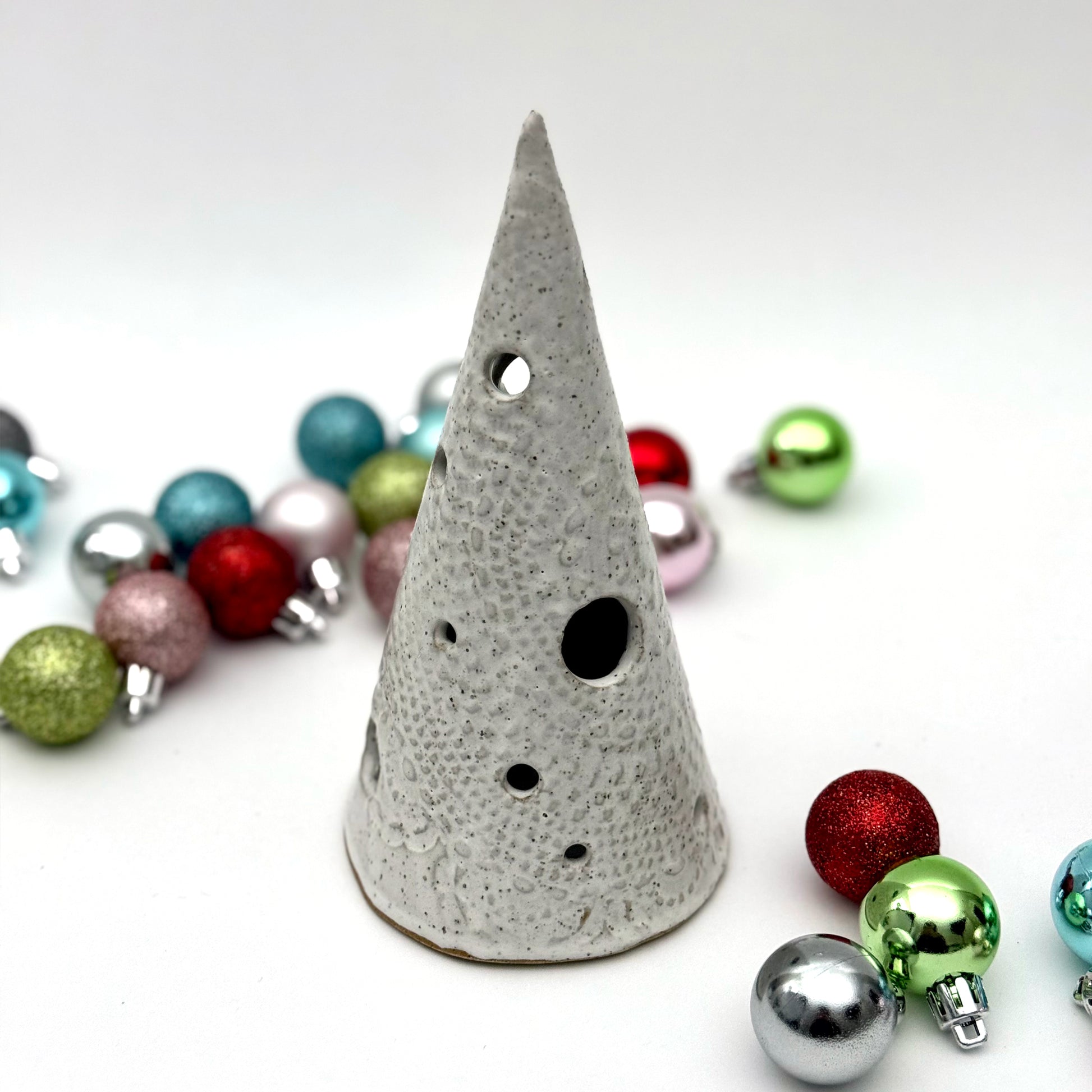 A white speckled ceramic tree-shaped luminary with light openings, styled among colorful holiday ornaments.