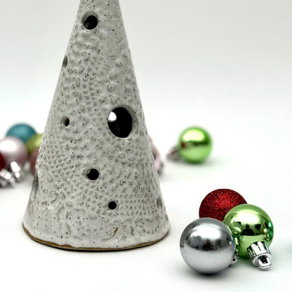 A white speckled ceramic tree-shaped luminary with light openings, styled among colorful holiday ornaments.