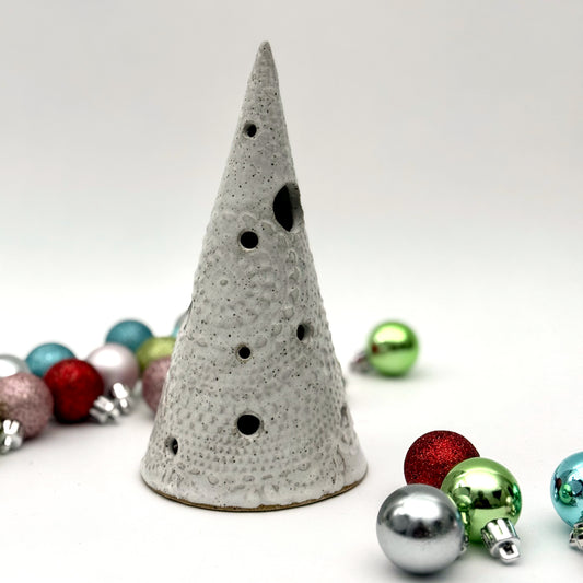 A white speckled ceramic tree-shaped luminary with light openings, styled among colorful holiday ornaments.
