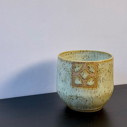 Handmade ceramic tumbler with a rounded shape and warm, yellow glaze. A square Empress breeze-block motif is inset on the front, revealing the natural clay and adding a mid-century geometric detail.