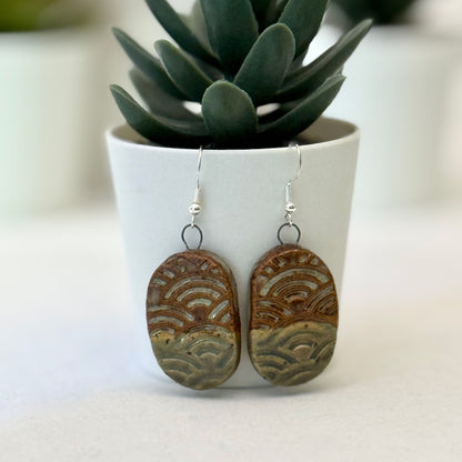 Pair of ceramic earrings with wave patterns in front of a potted plant.