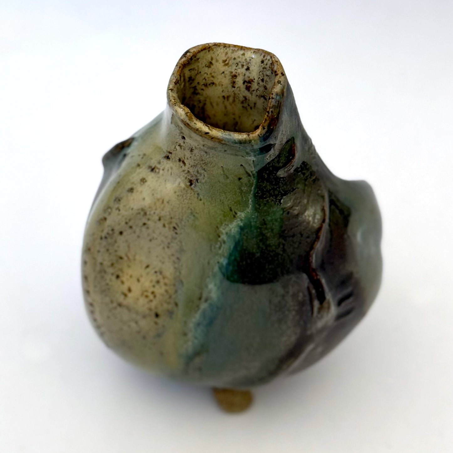 Handbuilt stoneware vase with a rounded organic form, textured surface impressions, and layered green and cream glaze. The sculptural piece stands on three small feet and has an earthy, natural finish.