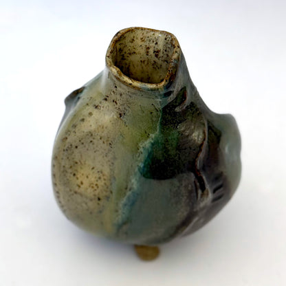 Handbuilt stoneware vase with a rounded organic form, textured surface impressions, and layered green and cream glaze. The sculptural piece stands on three small feet and has an earthy, natural finish.