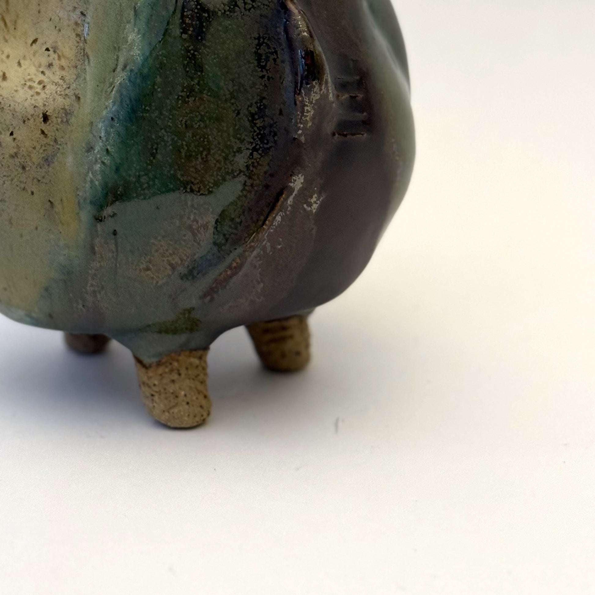 Handbuilt stoneware vase with a rounded organic form, textured surface impressions, and layered green and cream glaze. The sculptural piece stands on three small feet and has an earthy, natural finish.