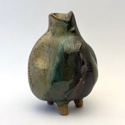 Handbuilt stoneware vase with a rounded organic form, textured surface impressions, and layered green and cream glaze. The sculptural piece stands on three small feet and has an earthy, natural finish.