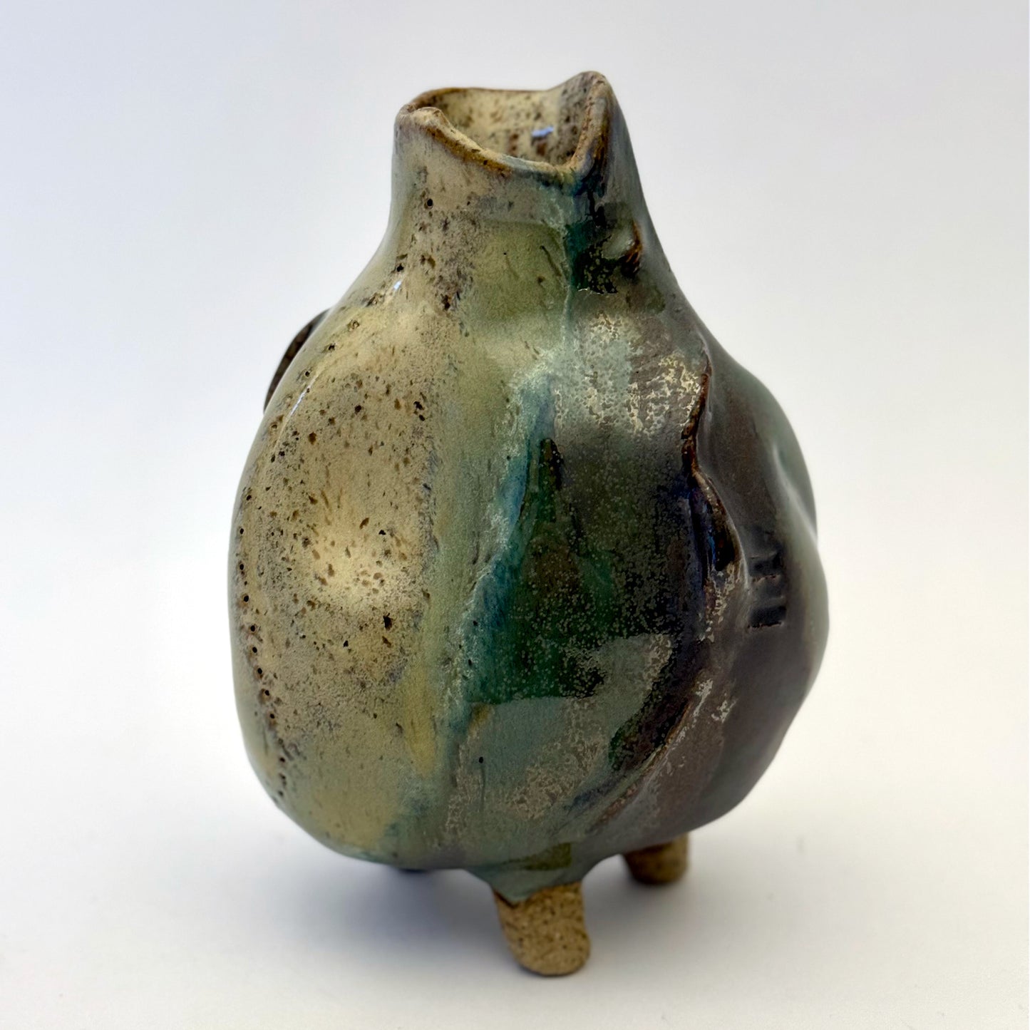 Handbuilt stoneware vase with a rounded organic form, textured surface impressions, and layered green and cream glaze. The sculptural piece stands on three small feet and has an earthy, natural finish.