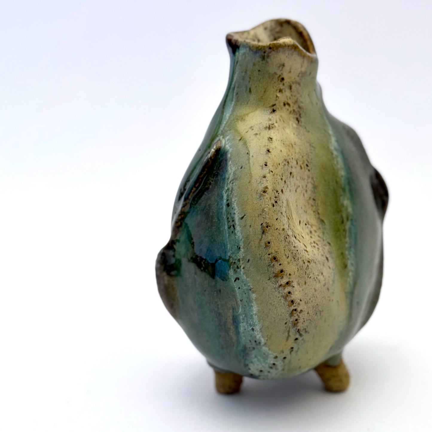 Handbuilt stoneware vase with a rounded organic form, textured surface impressions, and layered green and cream glaze. The sculptural piece stands on three small feet and has an earthy, natural finish.