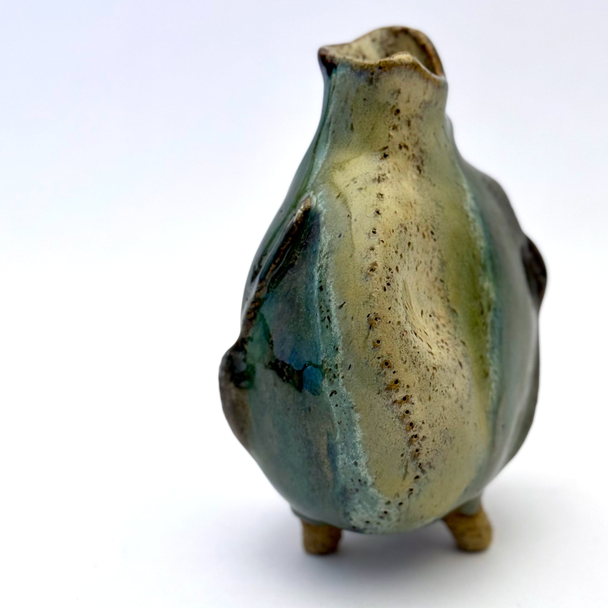 Handbuilt stoneware vase with a rounded organic form, textured surface impressions, and layered green and cream glaze. The sculptural piece stands on three small feet and has an earthy, natural finish.