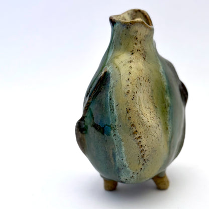 Handbuilt stoneware vase with a rounded organic form, textured surface impressions, and layered green and cream glaze. The sculptural piece stands on three small feet and has an earthy, natural finish.
