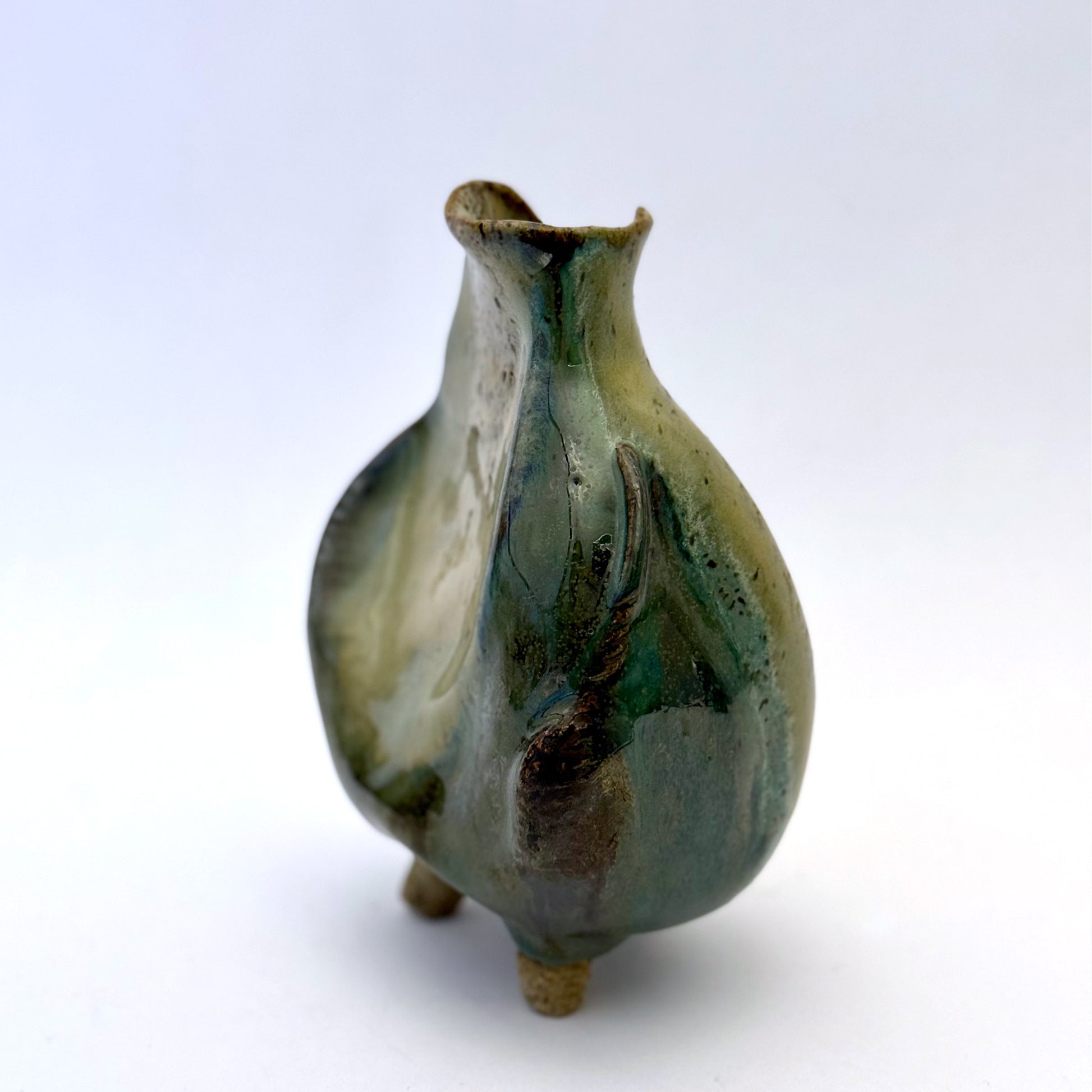 Handbuilt stoneware vase with a rounded organic form, textured surface impressions, and layered green and cream glaze. The sculptural piece stands on three small feet and has an earthy, natural finish.