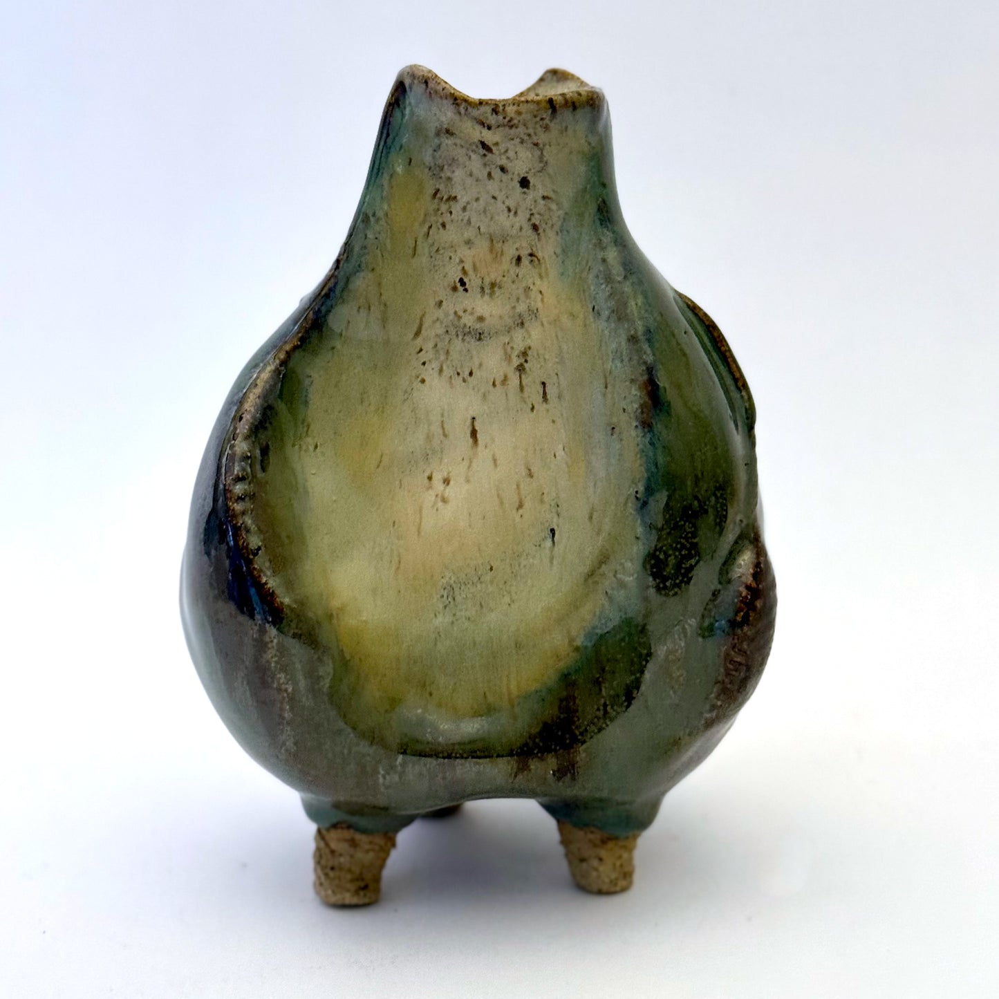 Handbuilt stoneware vase with a rounded organic form, textured surface impressions, and layered green and cream glaze. The sculptural piece stands on three small feet and has an earthy, natural finish.