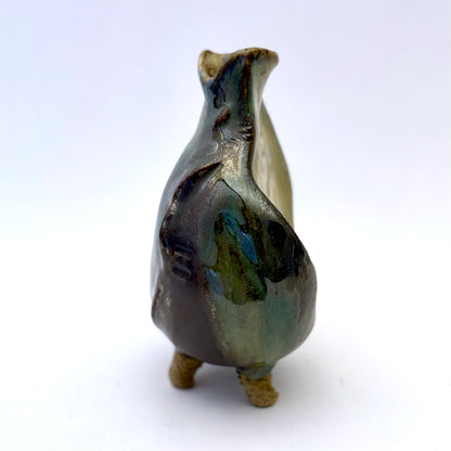 Handbuilt stoneware vase with a rounded organic form, textured surface impressions, and layered green and cream glaze. The sculptural piece stands on three small feet and has an earthy, natural finish.