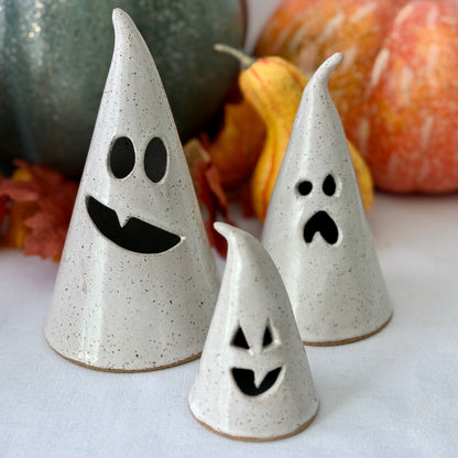 Three ceramic ghost luminaries with different facial expressions on a white surface with pumpkins in the background.