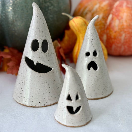 Three ceramic ghost luminaries with different facial expressions on a white surface with pumpkins in the background.