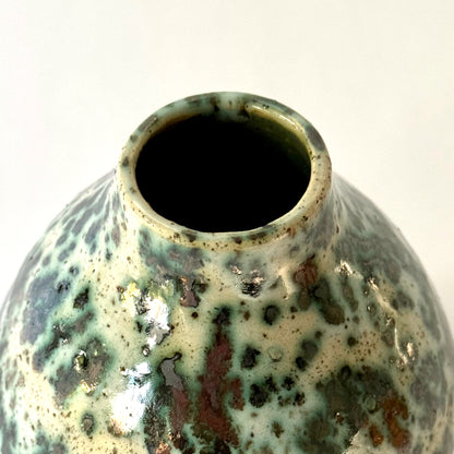 Close up of small handmade pottery vase with a rounded shape and narrow opening, finished in a green jasper glaze with varied green tones and irregular speckled patterning across the surface.
