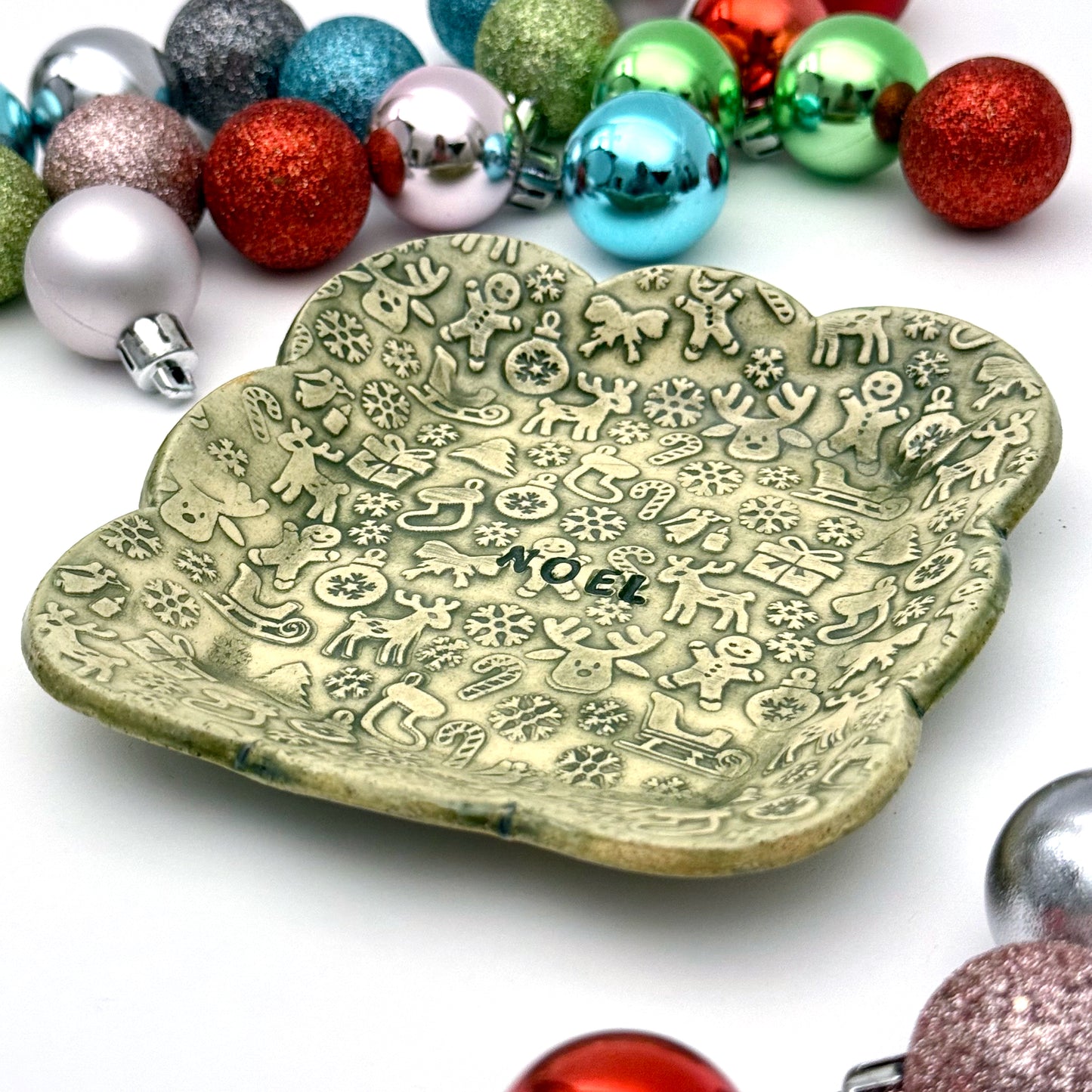 A green scalloped ceramic trinket dish featuring an all-over pattern of embossed Christmas symbols and the word “NOEL” in the middle, styled with shiny ornaments.