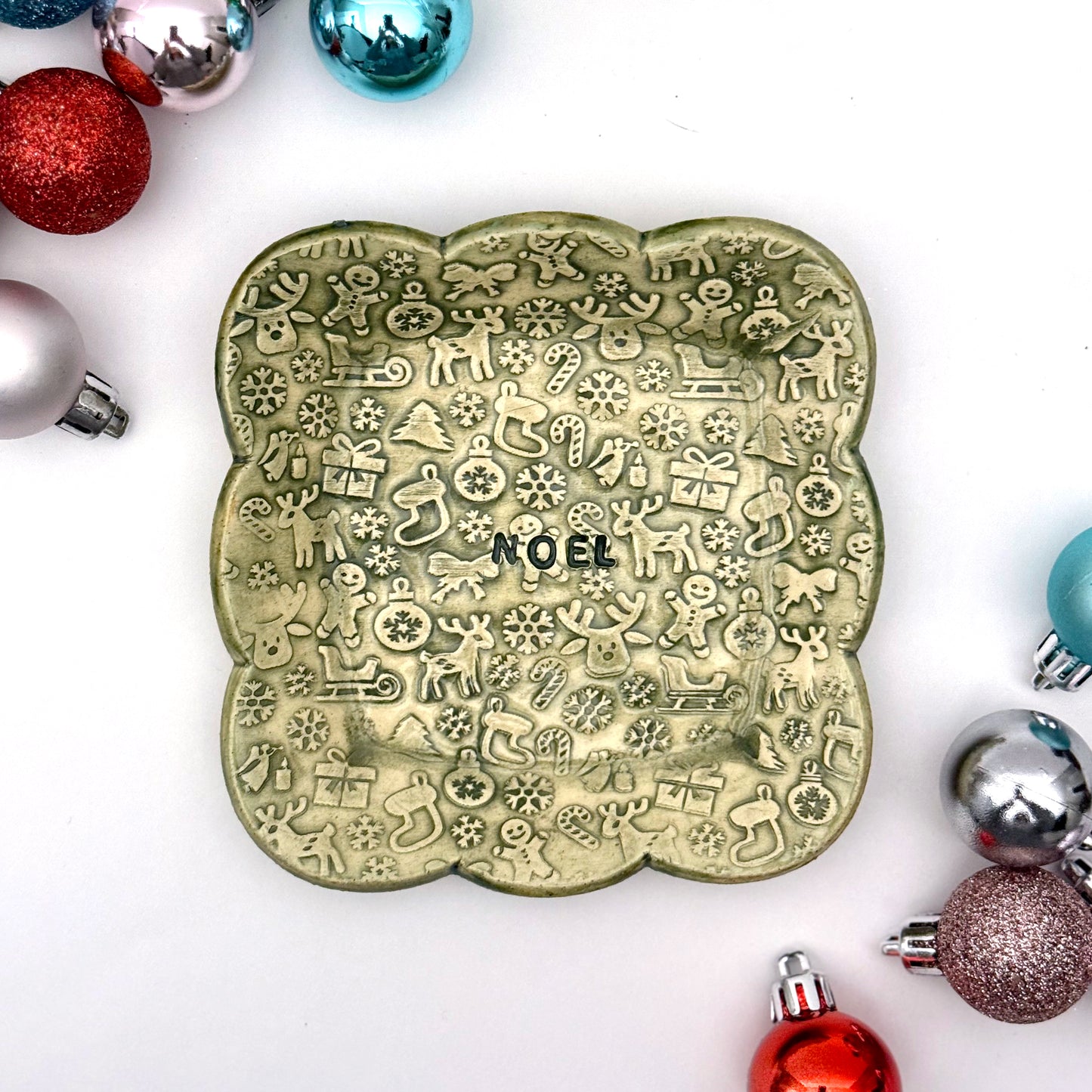 A green scalloped ceramic trinket dish featuring an all-over pattern of embossed Christmas symbols and the word “NOEL” in the middle, styled with shiny ornaments.