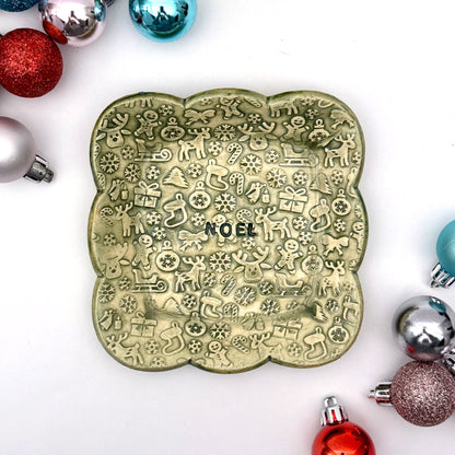 A green scalloped ceramic trinket dish featuring an all-over pattern of embossed Christmas symbols and the word “NOEL” in the middle, styled with shiny ornaments.