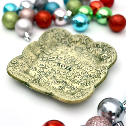 A green scalloped ceramic trinket dish featuring an all-over pattern of embossed Christmas symbols and the word “NOEL” in the middle, styled with shiny ornaments.