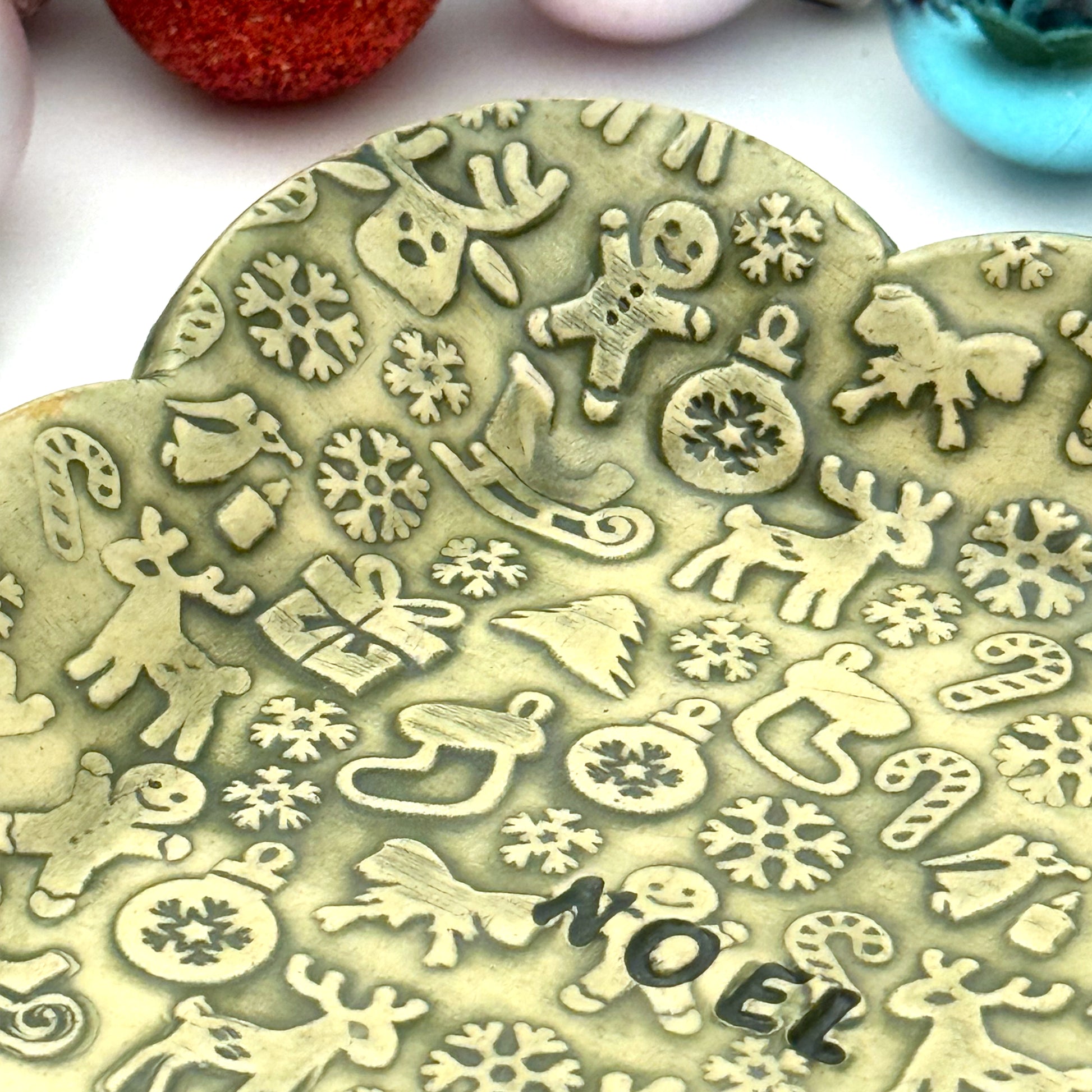 A green scalloped ceramic trinket dish featuring an all-over pattern of embossed Christmas symbols and the word “NOEL” in the middle, styled with shiny ornaments.