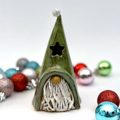 A ceramic gnome luminary with a green hat, star-shaped window, speckled white beard, and round nose, styled with Christmas ornaments.