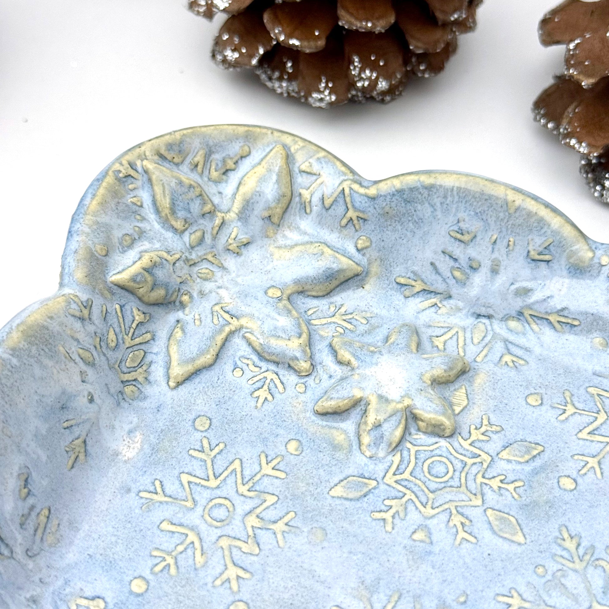 Close-up of A handmade blue ceramic dish with scalloped edges and raised snowflake patterns where the glaze breaks to reveal white clay, shown with decorative pinecones.