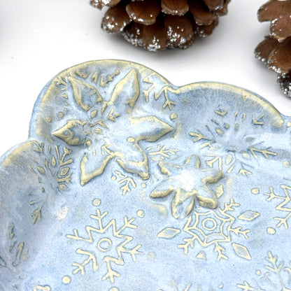 Close-up of A handmade blue ceramic dish with scalloped edges and raised snowflake patterns where the glaze breaks to reveal white clay, shown with decorative pinecones.