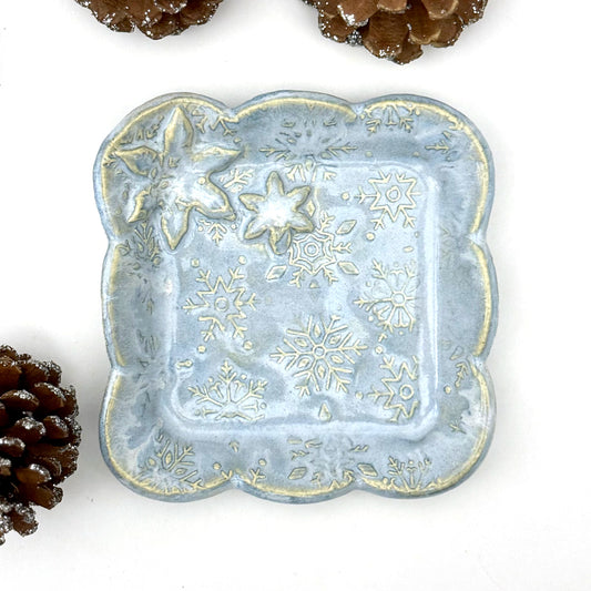 A handmade blue ceramic dish with scalloped edges and raised snowflake patterns where the glaze breaks to reveal white clay, shown with decorative pinecones.