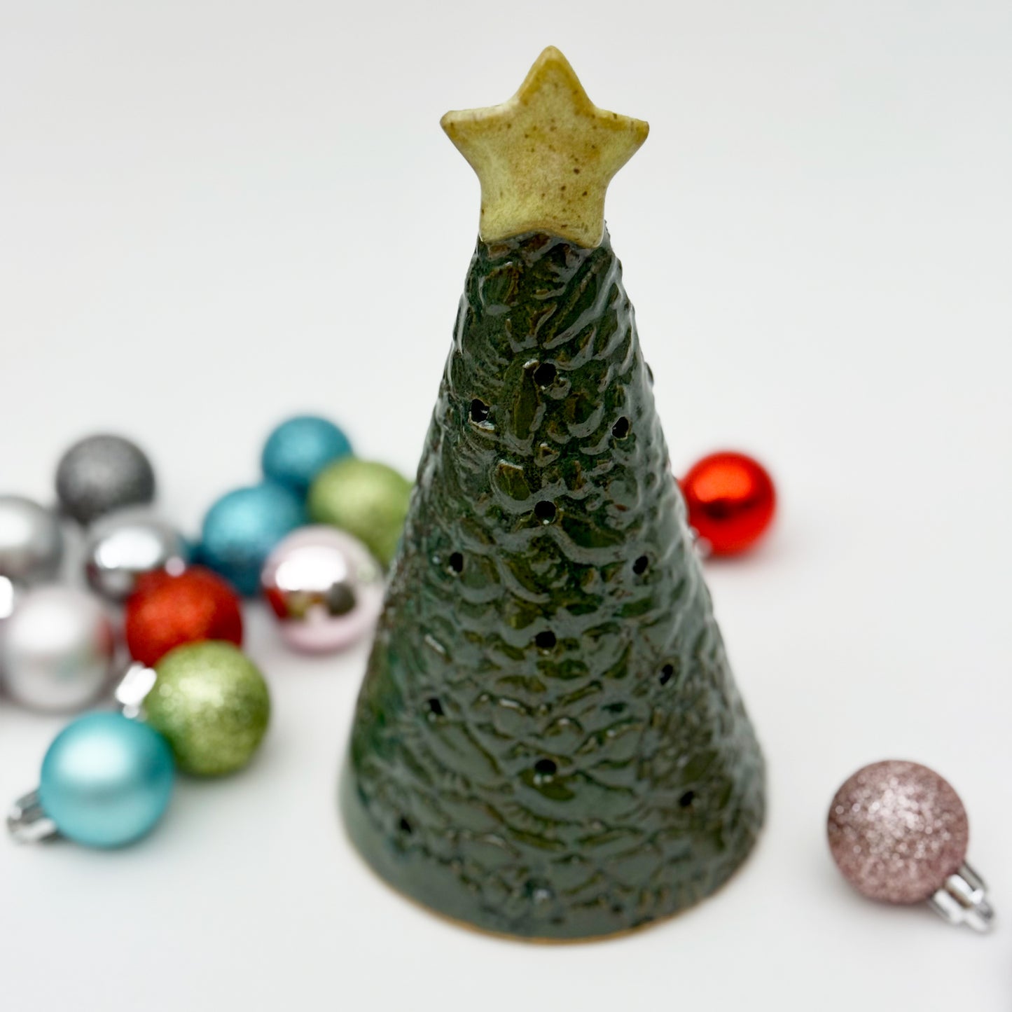 Green ceramic Christmas tree luminary with star on top, surrounded by colorful ornaments on a white background