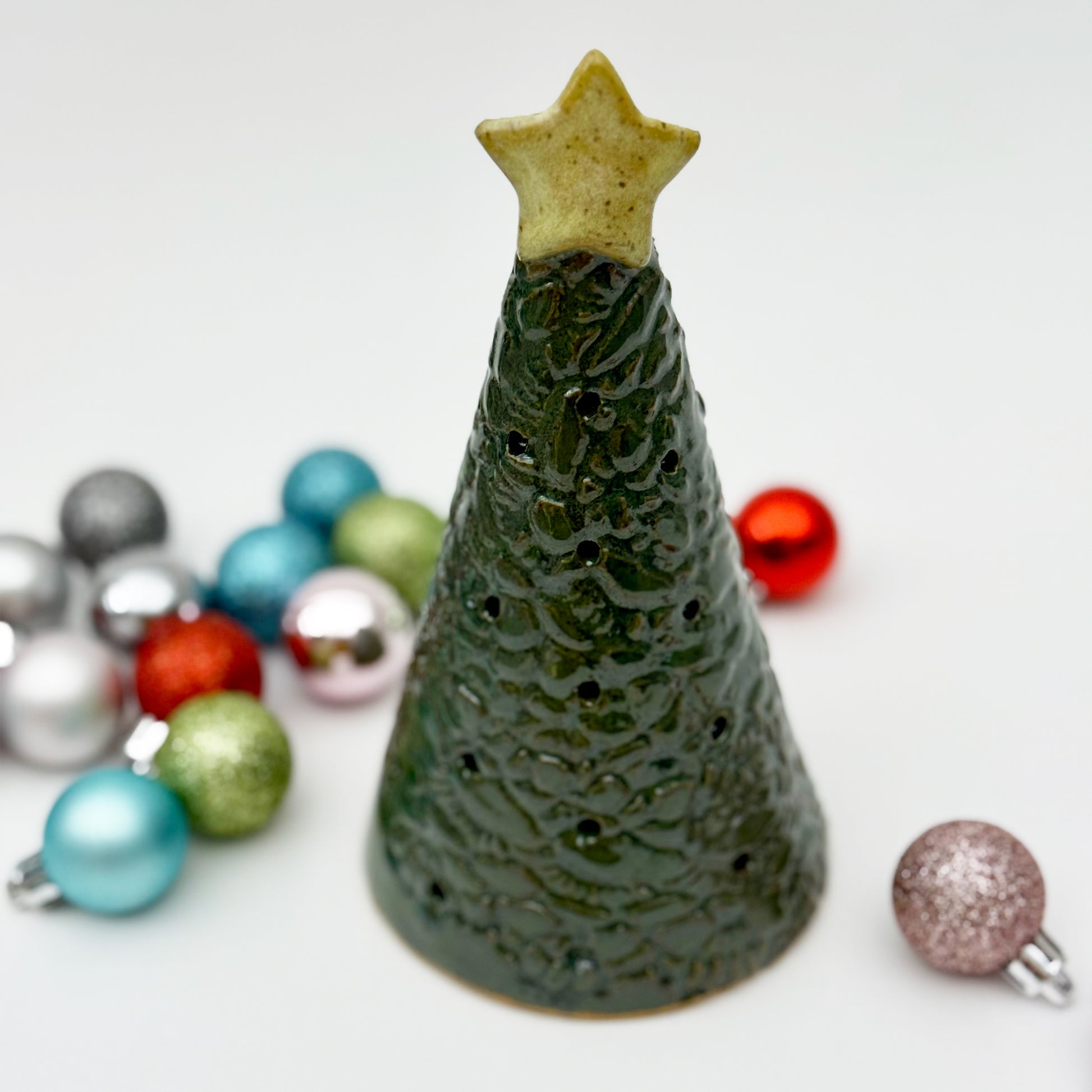 Green ceramic Christmas tree luminary with star on top, surrounded by colorful ornaments on a white background