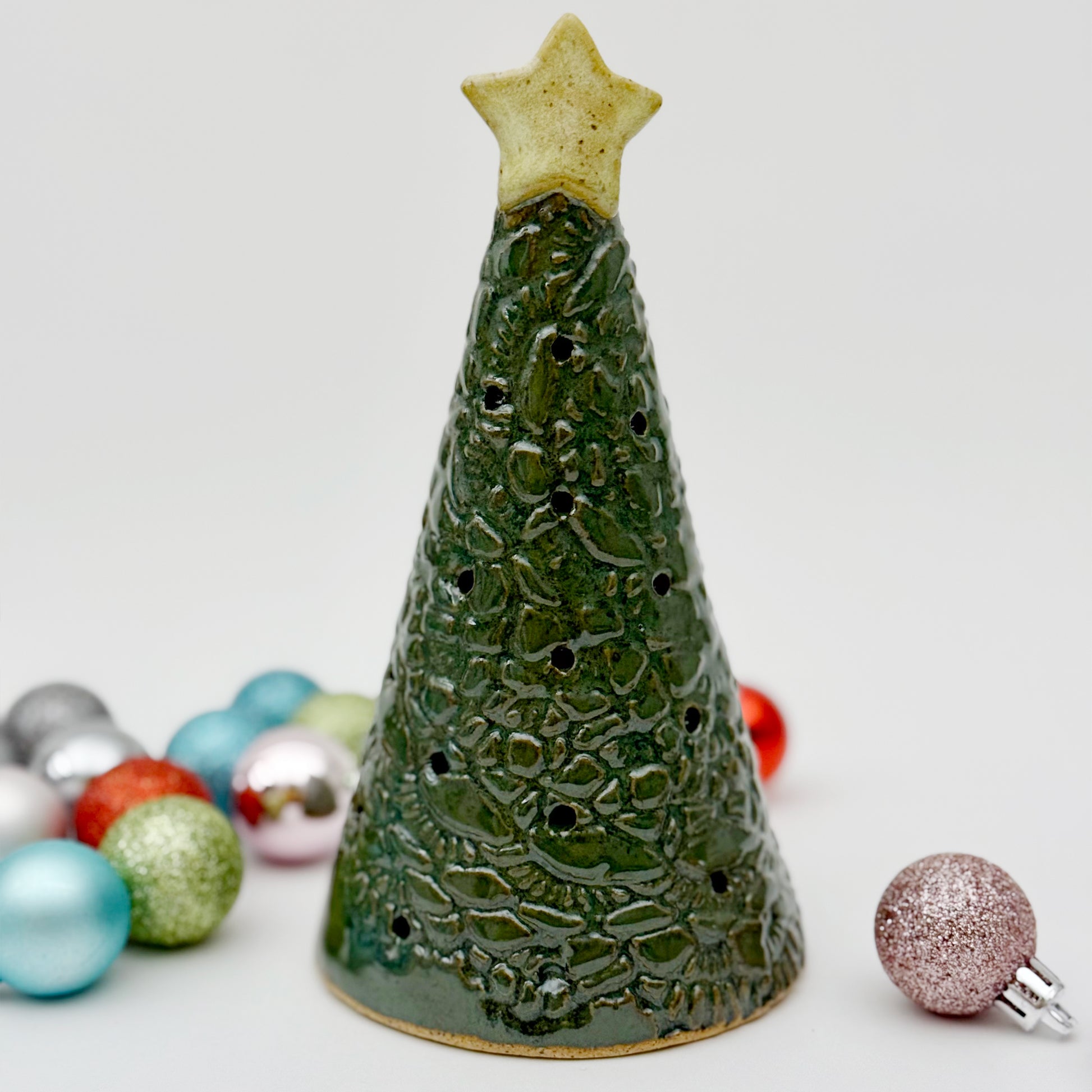 Ceramic Christmas tree luminsary with star on top, surrounded by colorful ornaments on a light background