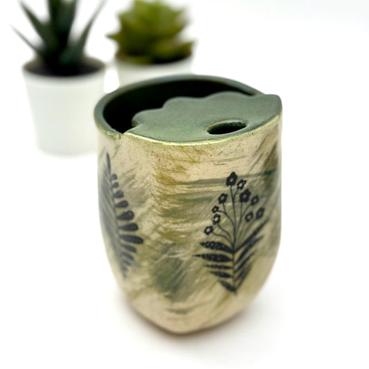 A handmade ceramic to-go cup in soft green and tan tones with black botanical designs. It includes a matching leaf-shaped lid with a steam hole. Blurred potted succulents sit in the background.