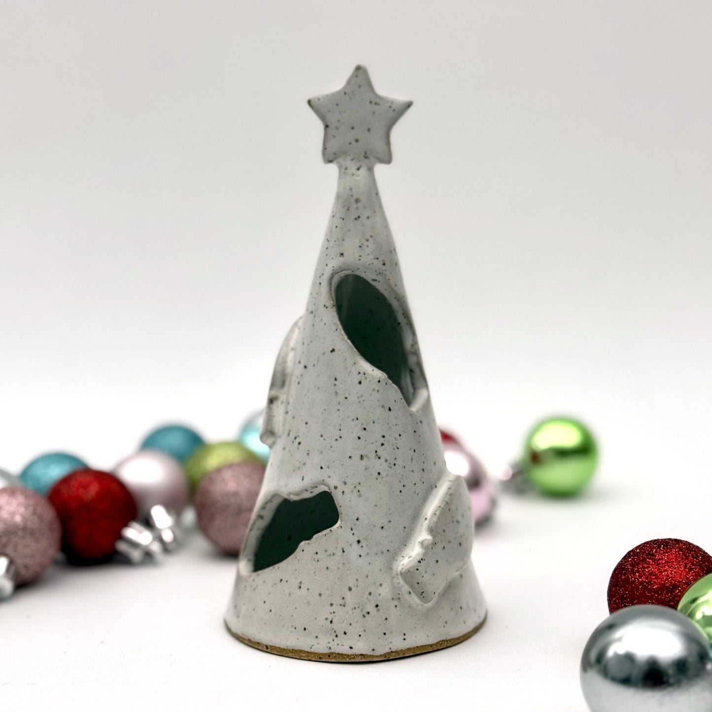 A white speckled ceramic tree luminary with cutout holiday light shapes and a star on top, surrounded by small colorful ornaments.