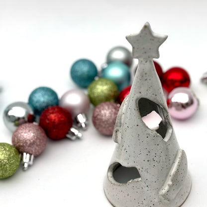 A white speckled ceramic tree luminary with cutout holiday light shapes and a star on top, surrounded by small colorful ornaments.